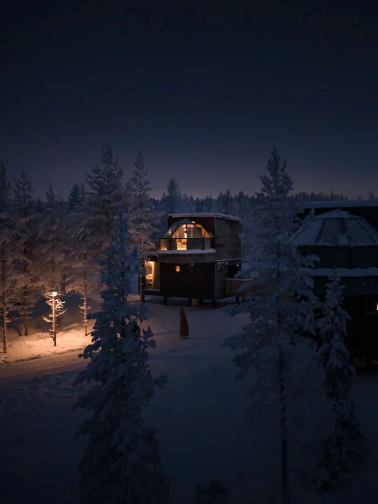 Warmly lit two-story glass igloo surrounded by snowy pine woods at night.